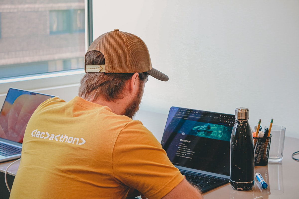 Developer in a baseball cap focused on coding across multiple laptop screens in a modern workspace
